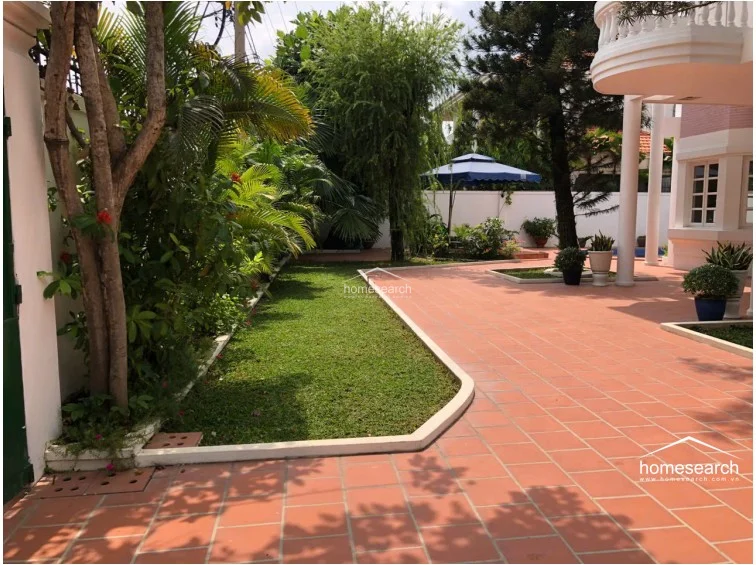 This expansive outdoor area features a well-maintained lawn bordered by lush tropical greenery and a large, terracotta-tiled patio perfect for lounging. The space connects seamlessly to the main residence, which showcases architectural columns and a balcony, offering a serene and private garden setting.