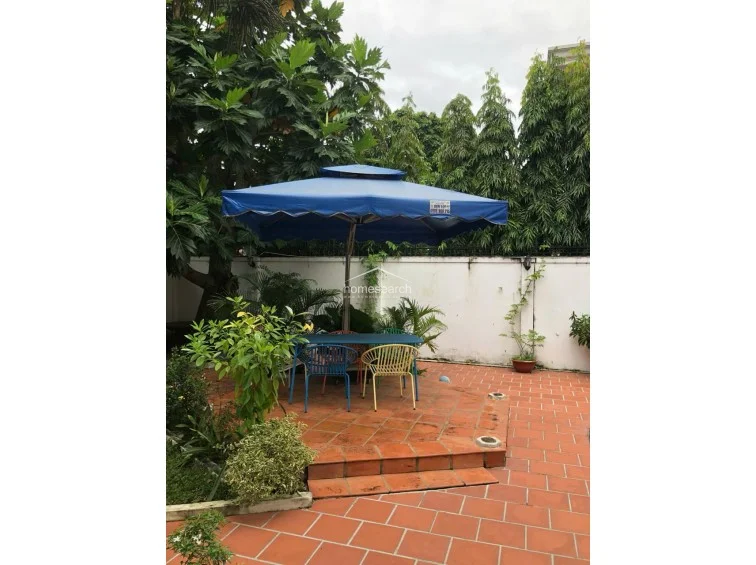 This outdoor patio area features terracotta-style tiled flooring and is shaded by a large, blue cantilever umbrella. The space is surrounded by lush greenery and a high privacy wall, creating a secluded retreat for outdoor dining or relaxation.