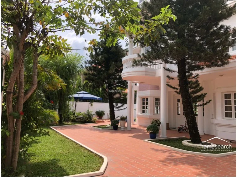 This exterior view showcases a well-maintained villa with a prominent white balcony, classical columns, and a terracotta-tiled terrace. The property is surrounded by mature trees and manicured garden beds, contributing to a serene and upscale residential atmosphere.