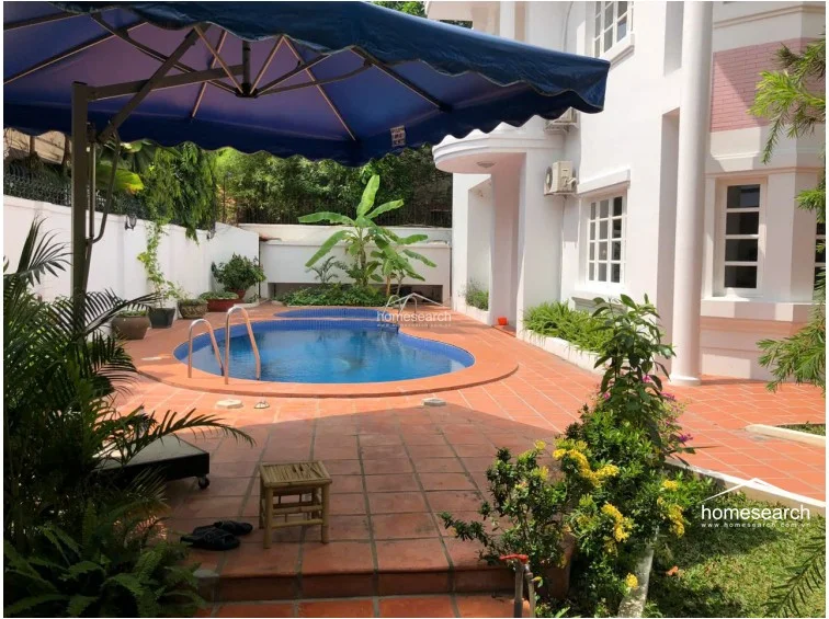 This outdoor area features a private, kidney-shaped swimming pool and a smaller circular plunge pool set into a terracotta-tiled deck. The space is shaded by a large blue umbrella and bordered by tropical plants and a white privacy wall, presenting a well-maintained and inviting recreational area.