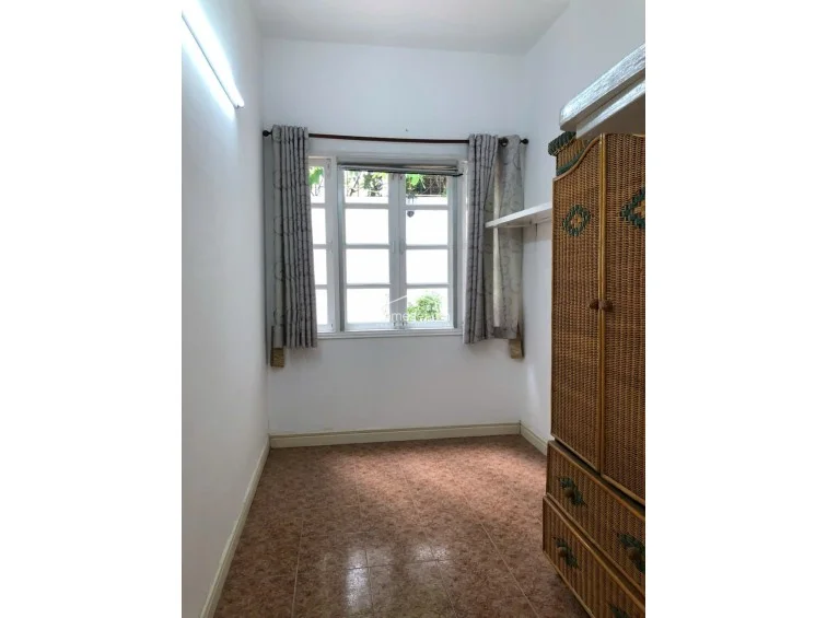 This smaller, functional room serves as a utility or storage space, featuring tiled flooring and a large, woven rattan wardrobe for storage. A window with light-filtering curtains provides natural light, and the space is finished with simple, neutral wall tones.