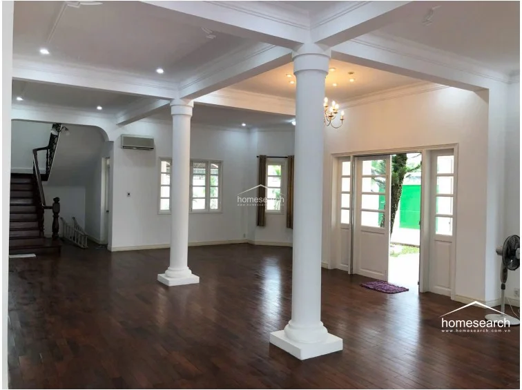 This spacious, open-concept living area features rich dark hardwood flooring and is anchored by prominent white structural columns. The room benefits from abundant natural light through multiple windows and glass-paned doors that provide a direct view of the outdoor garden space.