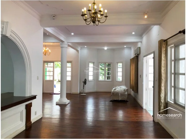 This bright living space showcases a classic architectural design with an elegant arched entryway and a traditional chandelier. The room maintains a consistent flow with dark wood flooring, white walls, and large windows that offer views of the exterior greenery.