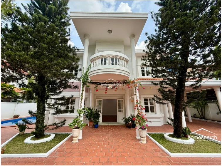 This image captures the grand entrance of the villa, emphasizing the symmetrical neoclassical design with its central balcony, white columns, and decorative bougainvillea vines. The front yard is neatly paved with terracotta tiles and features symmetrical garden beds with mature trees, creating a welcoming and elegant arrival experience. The structure appears to be in excellent condition, reflecting a classic architectural style.
