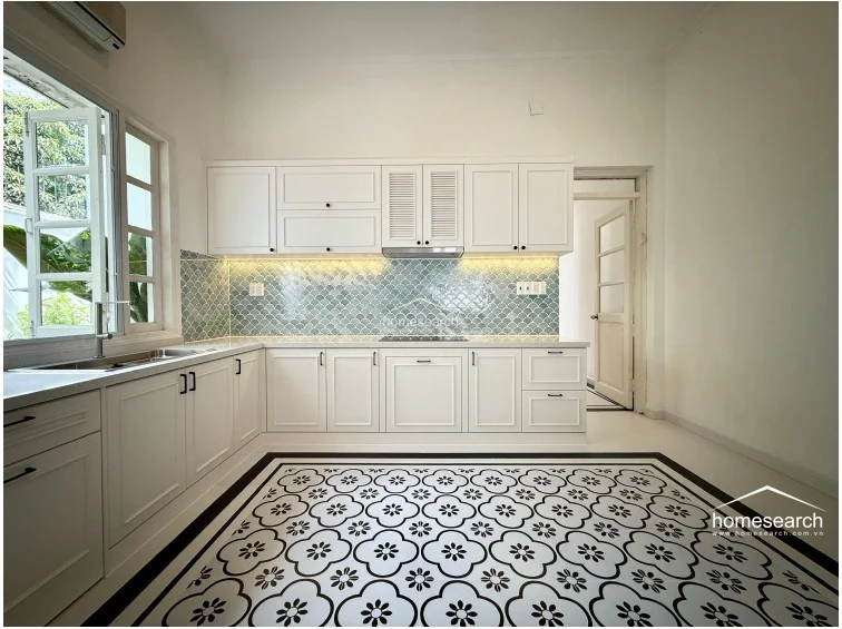 This bright, modern kitchen features white shaker-style cabinetry, high-quality stone countertops, and a unique, light-blue fish-scale tile backsplash. The floor is covered in a decorative, patterned tile that adds character, while the layout provides efficient workspace and direct access to an adjacent room. It is equipped with modern fixtures and recessed lighting, presenting a clean and functional culinary space.