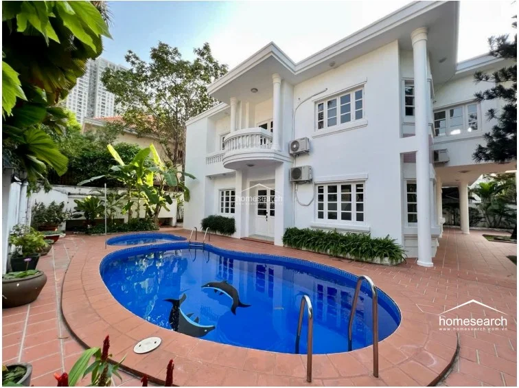 This exterior view showcases a private swimming pool set within a paved terrace area, featuring playful dolphin motifs on the pool floor. The two-story white villa displays neoclassical architectural elements, including prominent columns and a curved balcony, while the surrounding garden provides lush greenery and privacy. The property appears well-maintained with exterior AC units mounted for each level.