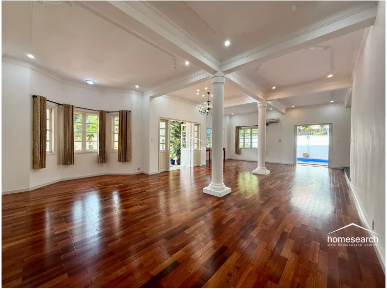 This expansive open-plan living and dining area features rich, polished hardwood flooring and is anchored by prominent white structural columns. The space is flooded with natural light from multiple windows dressed in neutral curtains, and it offers a seamless flow toward a rear terrace and pool area. The ceiling is accented with elegant crown molding, and the overall condition appears pristine and well-maintained.