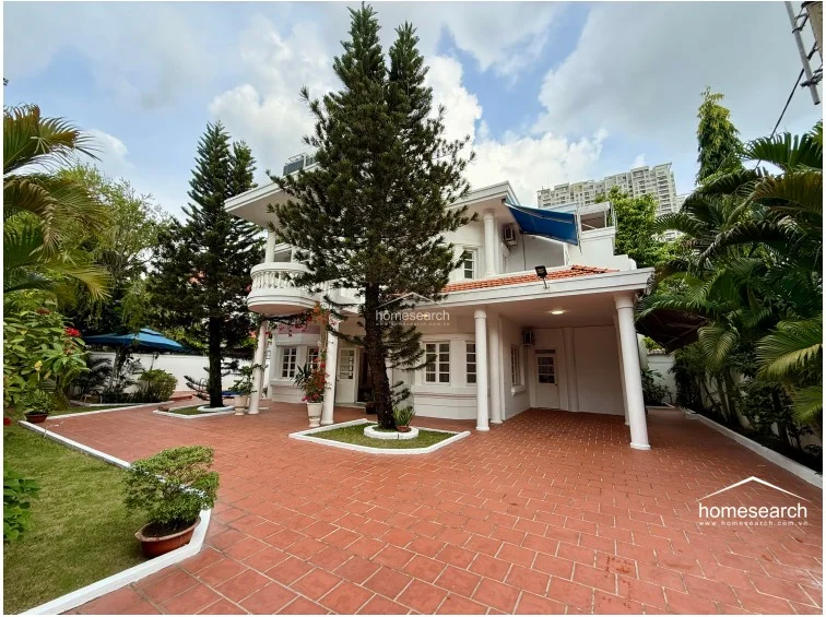This wide-angle shot of the villa exterior highlights the expansive paved driveway and front garden, which features mature pine trees and manicured landscaping. The property includes a covered carport area and a grand entrance framed by classical columns and a balcony above. The overall condition of the white facade and tiled outdoor areas suggests a high level of maintenance and curb appeal.