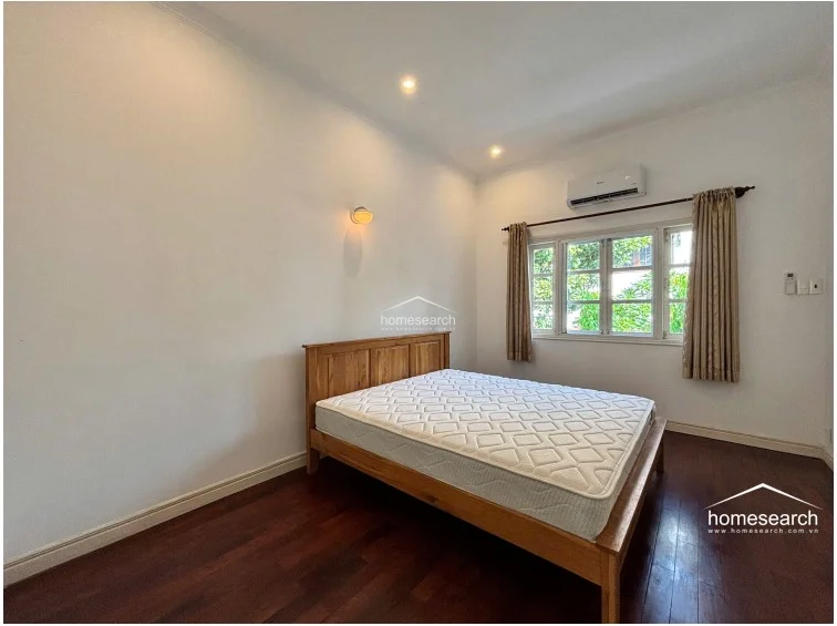This bedroom is finished with clean white walls and warm, dark wood flooring, creating a simple and inviting aesthetic. It is equipped with a wall-mounted air conditioning unit and a multi-pane window that provides natural light and views of the surrounding greenery.