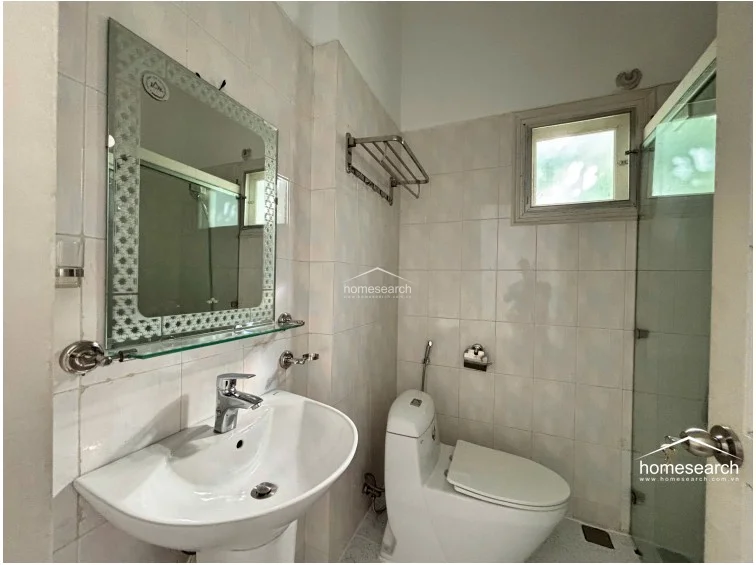 This functional bathroom is finished with light-colored wall tiles and a speckled terrazzo-style floor, creating a bright and clean atmosphere. It is equipped with a modern pedestal sink, a glass-enclosed shower, and a window that provides natural ventilation and light.