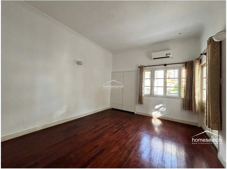 This bedroom features warm-toned hardwood flooring and crisp white walls, creating a clean and minimalist aesthetic. It is equipped with a wall-mounted AC unit and a built-in white wardrobe, while large windows with neutral curtains allow for ample natural light and a pleasant neighborhood view.