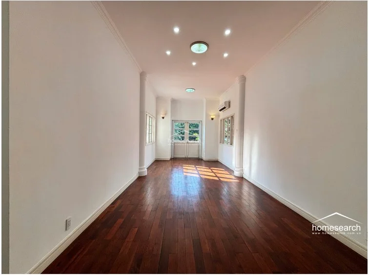 This long, gallery-style hallway or living space is finished with rich, dark hardwood flooring and white walls, providing a bright and airy atmosphere. The room is illuminated by recessed ceiling lights and wall sconces, with architectural columns framing the transition to an exterior doorway that leads to the outdoors.