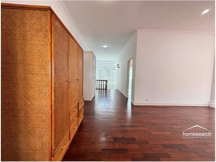 This spacious room or hallway features a large, vintage-style rattan wardrobe that adds a warm, organic texture to the minimalist white interior. The space is finished with high-quality hardwood flooring and leads into a bright, open landing area characterized by decorative architectural brickwork and a wooden staircase railing.