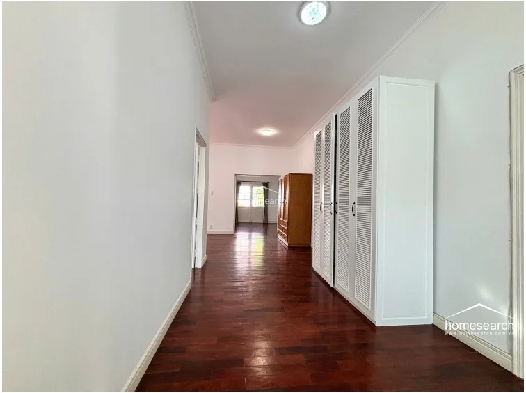 This interior hallway or bedroom transition space features dark, polished wood flooring and bright white walls, providing a clean and classic aesthetic. A large, white, louvered-door wardrobe offers substantial storage, while the layout flows directly into an adjacent room with a view of the exterior through glass doors.