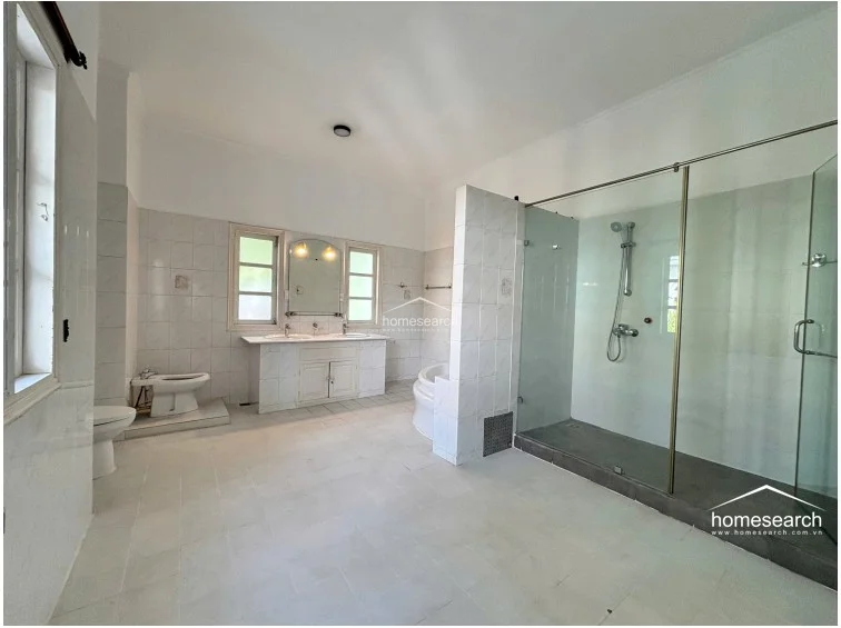 This expansive bathroom features a clean, white-tiled aesthetic with a dedicated glass-enclosed shower area and a separate soaking tub. The room is well-lit by natural light from two windows and includes a double-sink vanity with built-in storage cabinets underneath.