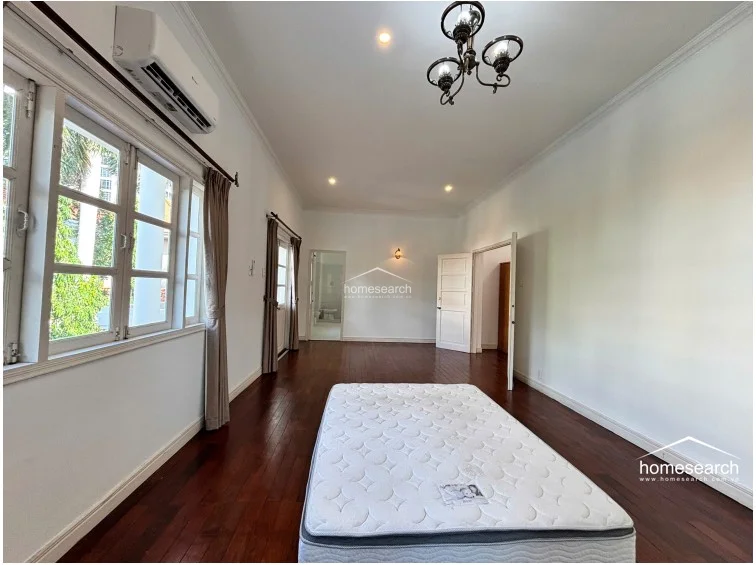 This spacious bedroom is finished with rich, dark wood flooring and neutral white walls, creating a bright and airy environment. It is equipped with a wall-mounted AC unit, a decorative chandelier, and large windows that allow for ample natural light, with an en-suite bathroom visible through an open doorway.