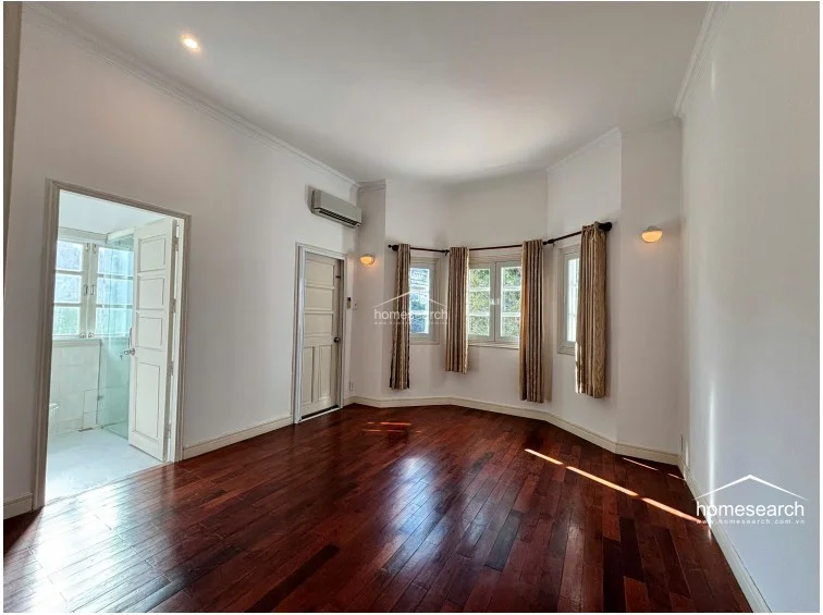 This bedroom features rich, dark wood flooring and crisp white walls, creating a clean and elegant aesthetic. The room is highlighted by a unique bay window area that invites natural light, complemented by an en-suite bathroom entrance and a wall-mounted AC unit.