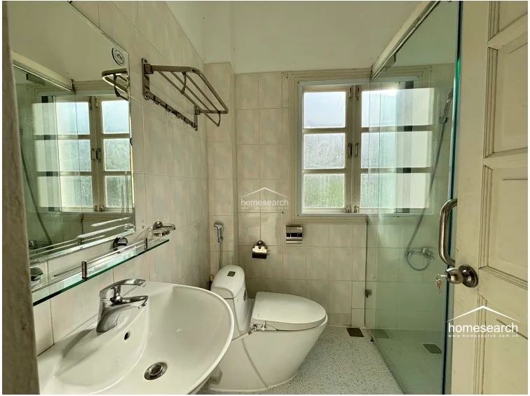 The bathroom is efficiently designed with a glass-enclosed shower stall, a modern pedestal sink, and a wall-mounted mirror with a glass shelf. It features light-colored tiled walls and floors, offering a clean, well-maintained space with a window for natural ventilation.