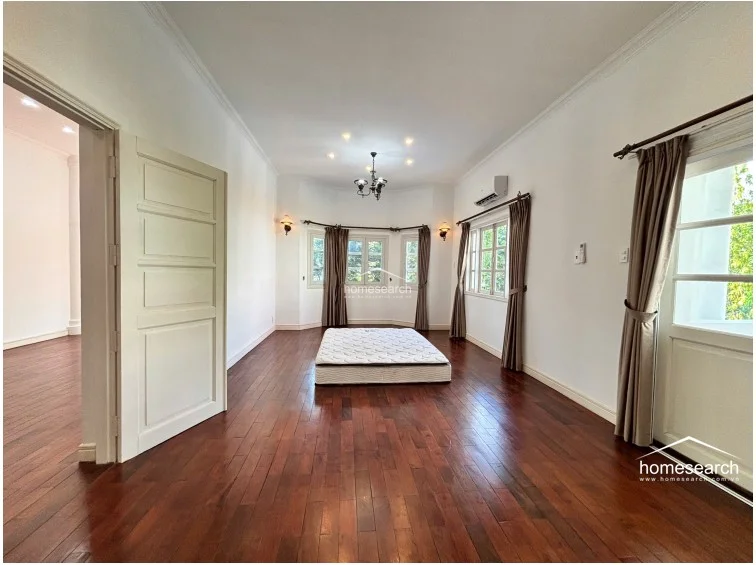 This spacious bedroom maintains a consistent design theme with dark wood flooring and white walls, centered around a minimalist mattress layout. The room includes a wall-mounted AC unit, classic wall sconces, and a large bay window arrangement that provides a bright, airy atmosphere.