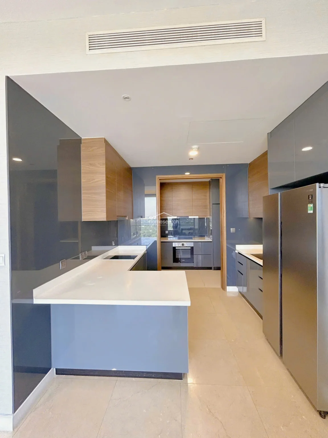 The modern kitchen is designed with a functional U-shaped layout, featuring high-gloss grey cabinetry paired with warm wood-tone upper cupboards and white stone countertops. It is well-equipped with integrated appliances, including a built-in oven and a large stainless steel refrigerator, all maintained in excellent condition.