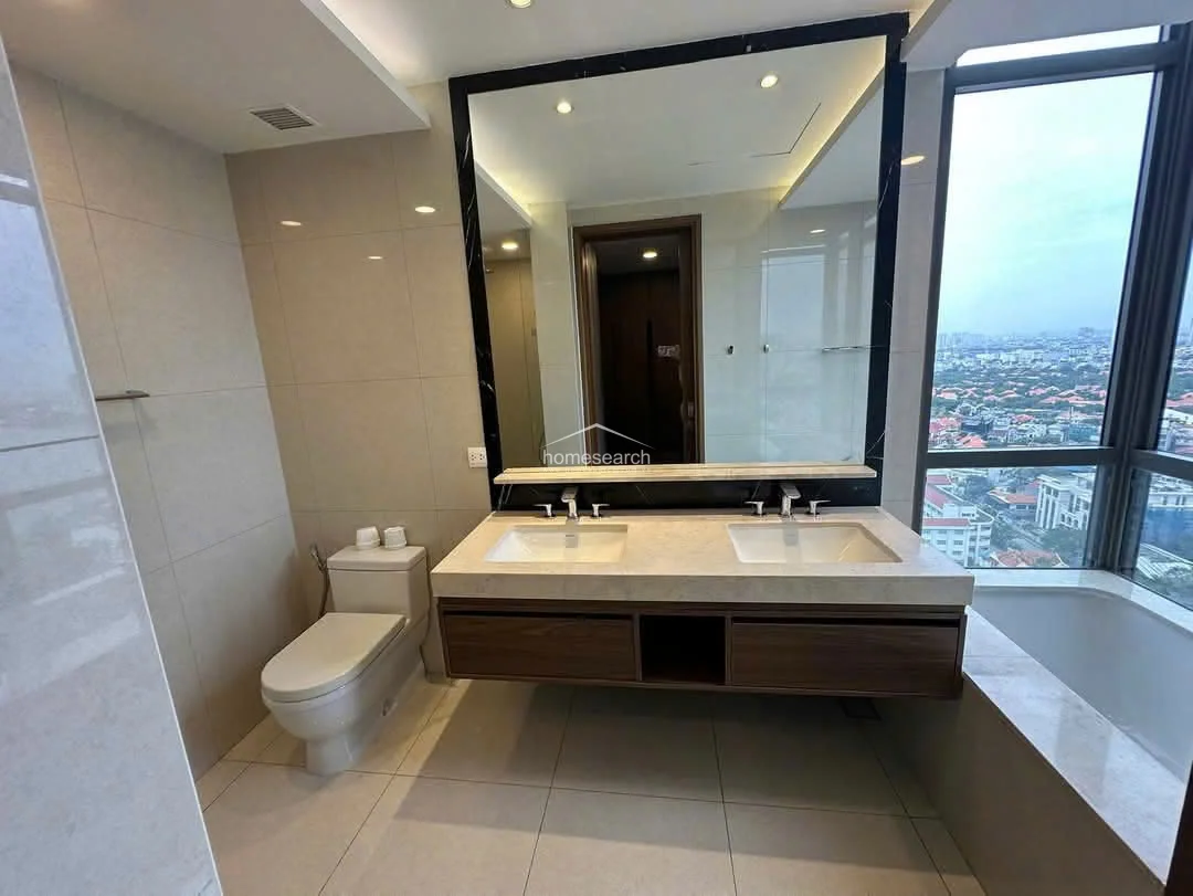 This primary bathroom is designed with a luxurious, modern aesthetic, featuring a dual-sink vanity with a floating wood cabinet and a large, dark-framed mirror. The space is finished with light-toned wall and floor tiles, and includes a deep soaking tub positioned directly against a window that offers impressive elevated views.