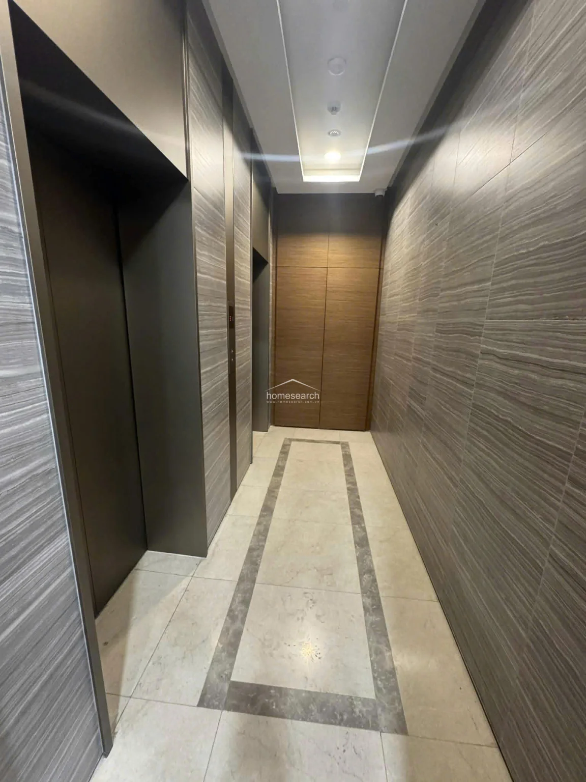 This elevator lobby features a sophisticated, modern aesthetic with floor-to-ceiling grey wood-grain paneling and recessed cove lighting. The flooring consists of large-format light tiles accented by a dark stone inlay border, creating a clean and high-end transition space.