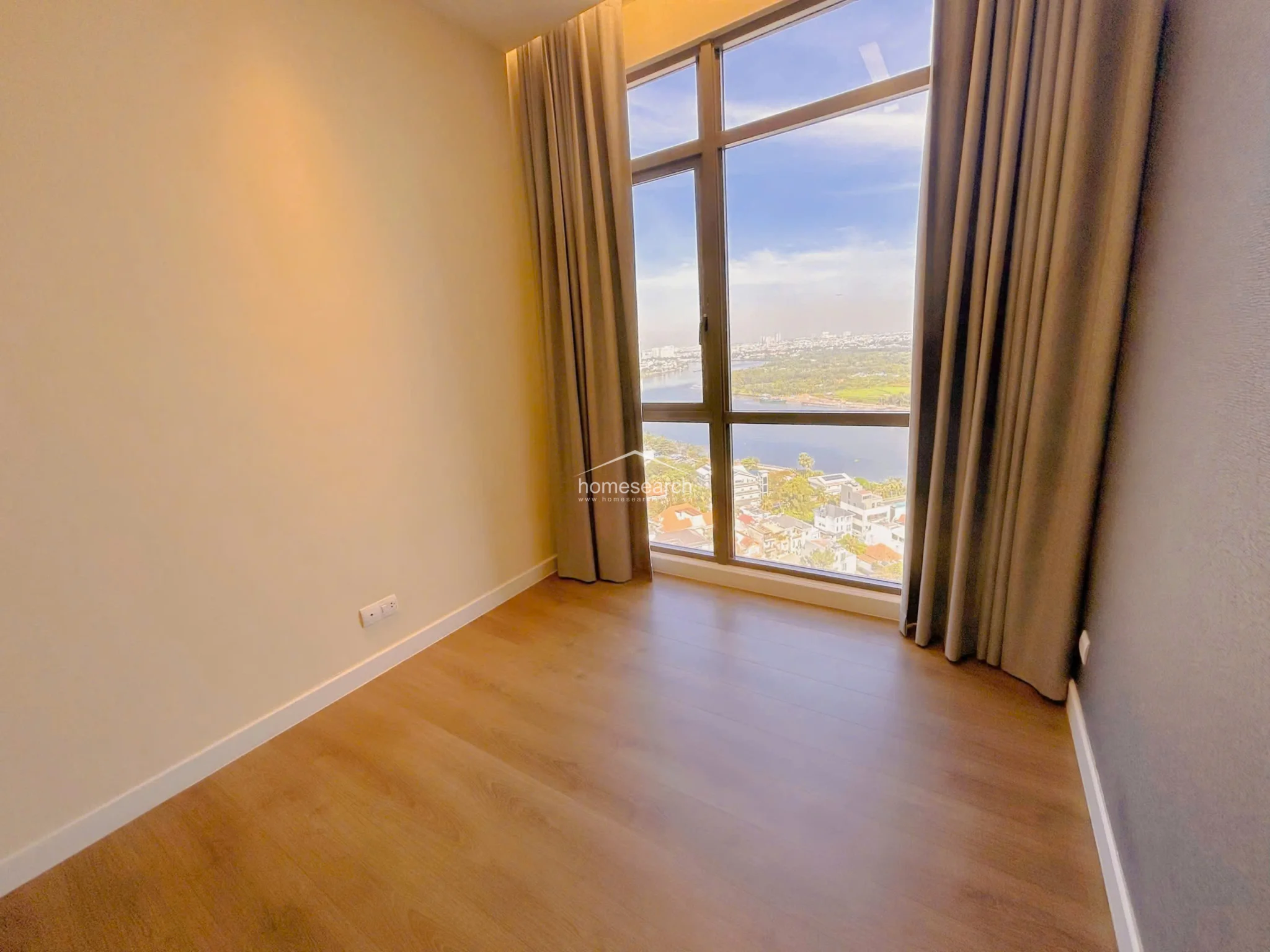 This bedroom offers a bright, minimalist atmosphere with warm wood-toned flooring and neutral wall finishes. The standout feature is the floor-to-ceiling window that provides an expansive, unobstructed view of the river and surrounding cityscape, complemented by simple floor-length curtains.