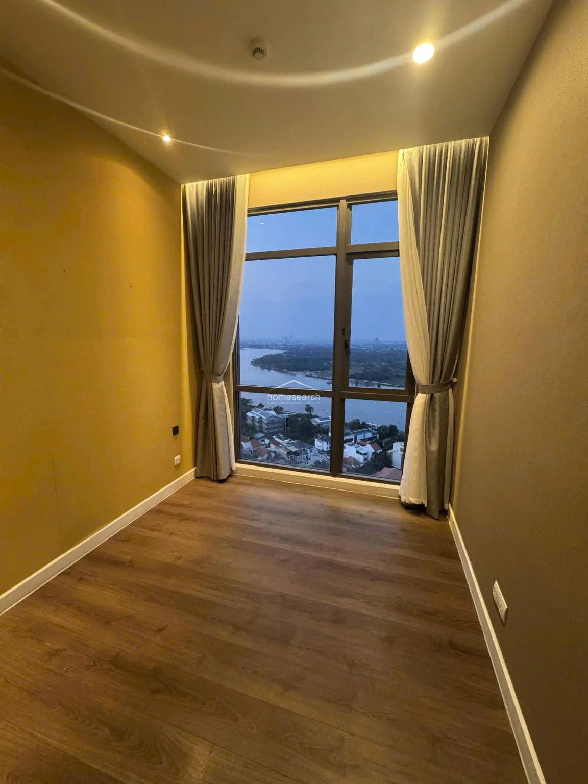 This bedroom is identical in layout to the previous image, featuring warm wood flooring and a neutral color palette that maximizes the sense of space. The large window serves as the focal point, framing a scenic river view and allowing for significant natural light, while the recessed ceiling lighting adds a soft, ambient glow.