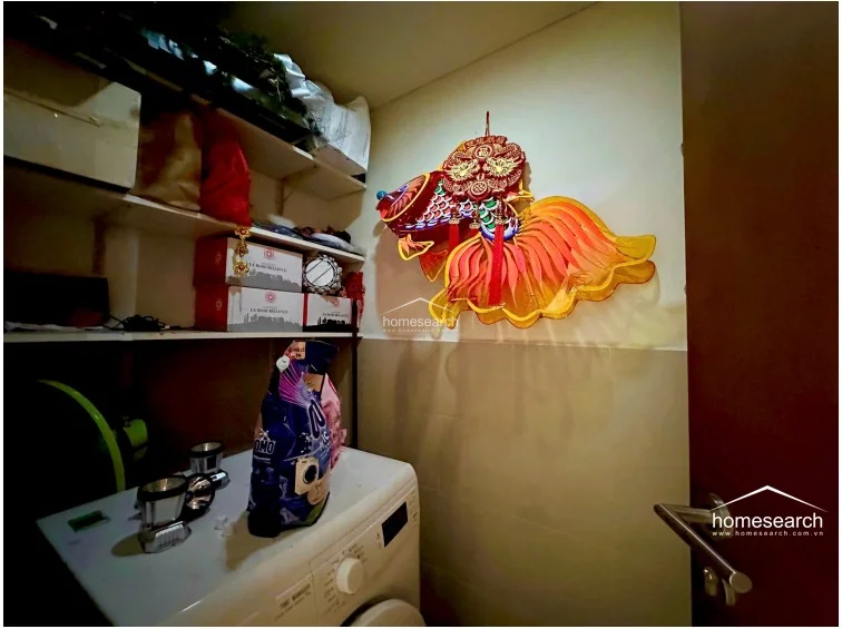 This utility or laundry area features built-in white shelving for storage and a front-loading washing machine. The space is finished with neutral-toned wall tiles and is decorated with a vibrant, traditional fish-shaped lantern, indicating a well-maintained, functional service zone.