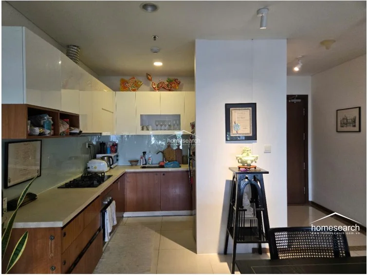 This perspective shows the kitchen’s integration into the main living area, highlighting the efficient use of space and the clean, contemporary design of the cabinetry. The inclusion of decorative elements, such as the traditional wooden stand and art pieces, adds a personalized touch to the otherwise minimalist and well-kept interior.