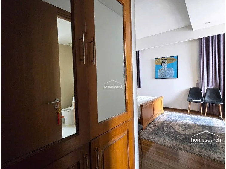 This bedroom features warm wood flooring and a classic wooden bed frame, complemented by a large, ornate area rug. A built-in wooden wardrobe with a mirrored panel provides storage and a view into the adjacent en-suite bathroom, which appears to have white tile finishes.