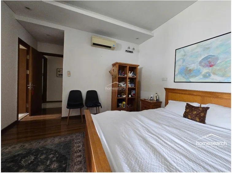 The bedroom is furnished with a wooden bed, matching nightstands, and an open-shelf storage unit, all set against neutral white walls. A wall-mounted AC unit is present, and the room is accented by two black modern chairs and a large, vibrant piece of wall art.