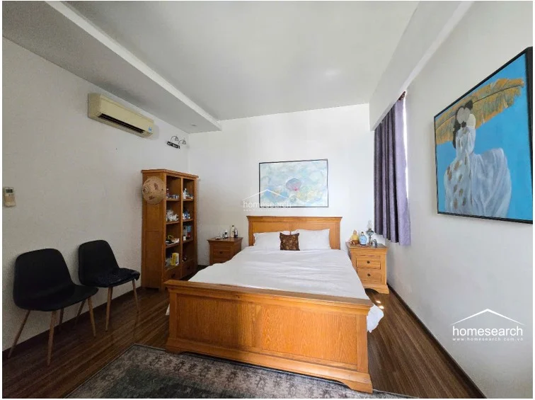 This view highlights the bedroom's layout, showing the bed centered between two nightstands and a tall wooden storage cabinet. The space is brightened by natural light filtered through purple curtains, while the wood flooring adds a cohesive, warm tone to the room.