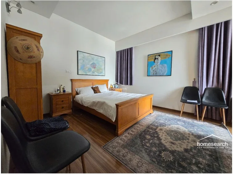 This perspective emphasizes the bedroom's furniture arrangement, including the two black chairs and the wooden wardrobe visible in the corner. The room maintains a clean, well-kept aesthetic with a balanced mix of traditional wooden furniture and contemporary art.