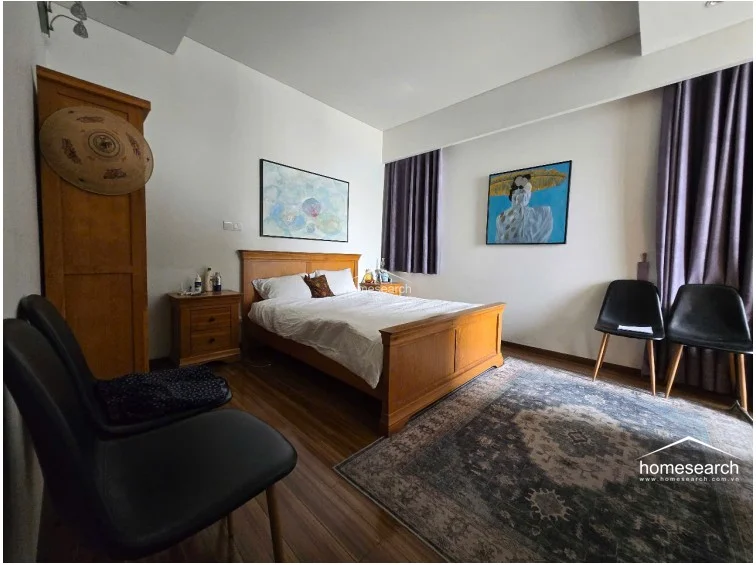 This primary bedroom features warm wood-look flooring and a classic wooden bed frame, complemented by a large area rug and two modern black accent chairs. The room is well-lit by natural light from a large window dressed in purple curtains, and it includes a tall wooden wardrobe for storage and decorative wall art.