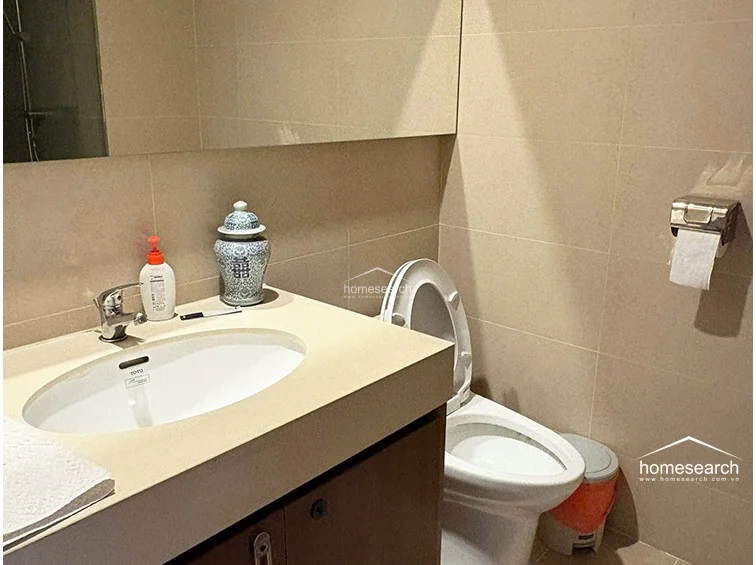 This bathroom is finished with neutral-toned wall and floor tiles, creating a clean and minimalist atmosphere. It is equipped with a Toto sink basin set into a vanity cabinet, a standard toilet, and a large wall-mounted mirror, all appearing to be in good, well-maintained condition.