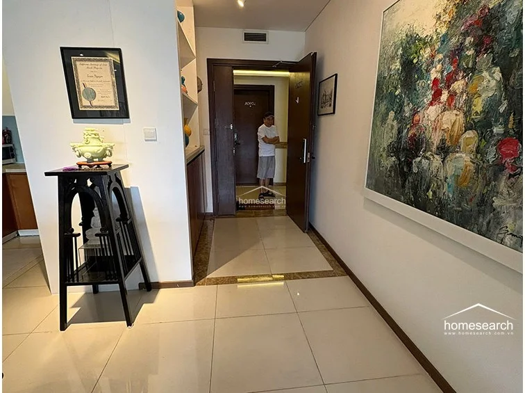 The entryway features light-colored floor tiles with a decorative border and leads directly into the main living space. A tall, dark wooden display stand sits near a built-in shelving unit, while a large, colorful abstract painting adds a focal point to the wall near the main entrance door.