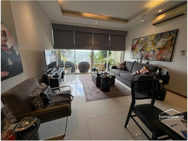 The living room is furnished with a comfortable grey sofa, a matching ottoman, and a dark wood media console, creating a cozy seating arrangement. Large Roman shades cover the balcony entrance, and the walls are adorned with large-scale contemporary artwork, highlighting a high level of maintenance and curated interior design.