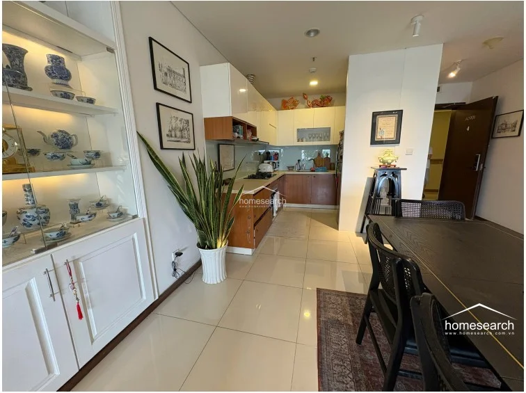 This view highlights an open-plan layout that seamlessly connects the kitchen, dining area, and living room, emphasizing a cohesive flow throughout the apartment. A large, illuminated glass-fronted display cabinet provides elegant storage, while the neutral white walls and polished tile flooring create a bright, airy atmosphere throughout the home.