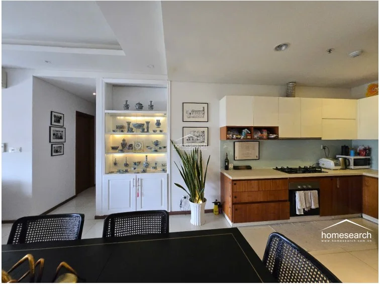 The kitchen features an L-shaped layout with warm wood-toned lower cabinets and sleek white upper cabinetry, complemented by a light-colored countertop. A prominent built-in display cabinet with integrated lighting showcases a collection of ceramics, while the adjacent dining area maintains a consistent, elegant design language throughout the home.