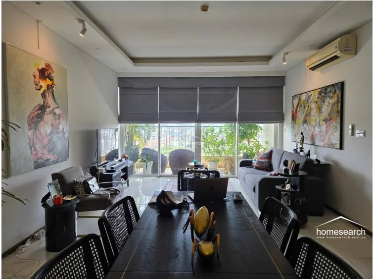 The living area is centered around a comfortable grey sofa and a dark wood dining set, illuminated by a mix of recessed ceiling lights and natural light filtering through the balcony doors. The space is decorated with large-scale artwork and features a wall-mounted AC unit, with the balcony providing a private outdoor extension that overlooks the surrounding cityscape.