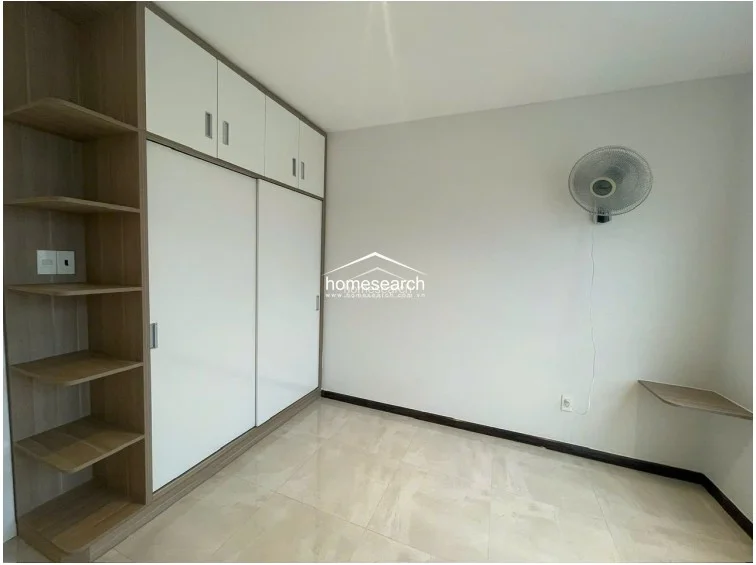 This bedroom is efficiently designed with a large, floor-to-ceiling wardrobe that combines white sliding doors with open wooden shelving for display. The room is finished with neutral walls and polished tile flooring, and it includes a wall-mounted fan and a small corner shelf for added convenience.