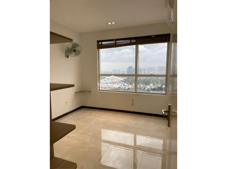 This bedroom or study area features large windows with a scenic river and city view, complemented by minimalist light-colored tile flooring. The room includes built-in wall shelving, a wall-mounted fan, and a bamboo-style window shade, maintaining a clean and functional aesthetic.