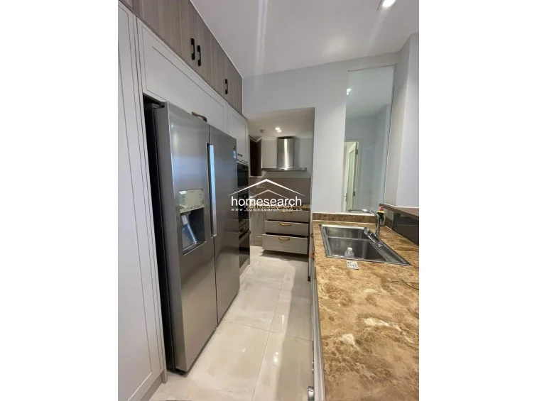 This modern kitchen is equipped with high-end stainless steel appliances, including a large double-door refrigerator and a built-in oven. It features rich brown marble countertops, sleek light-wood cabinetry for ample storage, and a clean, minimalist aesthetic with recessed lighting.