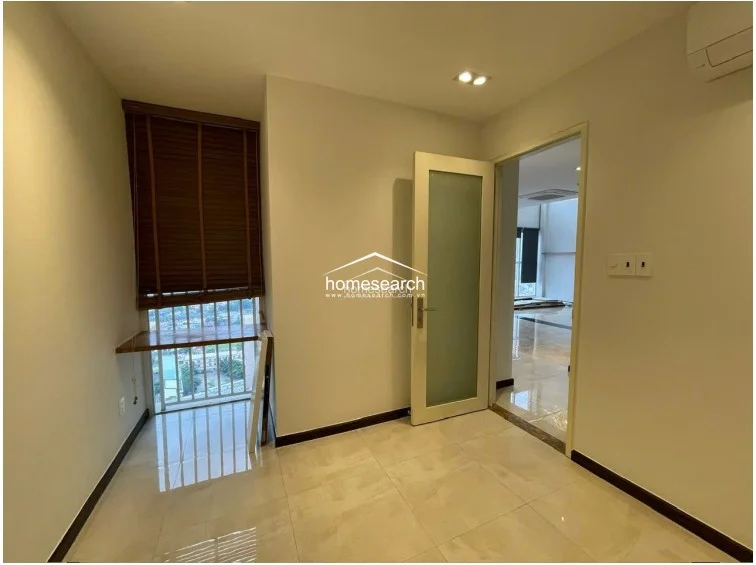 This small room serves as a versatile study or home office, featuring a built-in wooden desk positioned directly under a window with dark wood blinds. The space is finished with neutral cream walls and light-colored tile flooring, and it connects directly to the main living area through a frosted glass door.