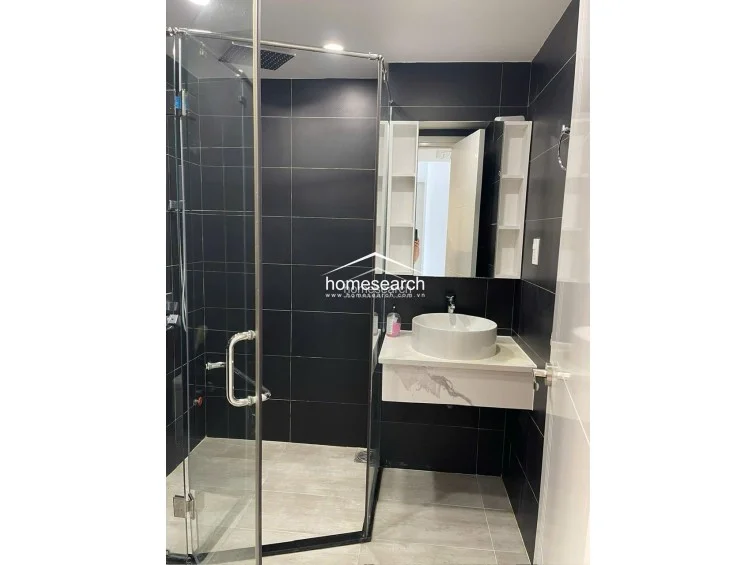 This contemporary bathroom utilizes a striking high-contrast design with matte black wall tiles and light-toned floor tiles. It is equipped with a glass-enclosed corner shower, a floating marble-topped vanity with a vessel sink, and built-in side shelving for organized storage. The overall condition is pristine, showcasing a sleek, minimalist aesthetic that maximizes the available space.