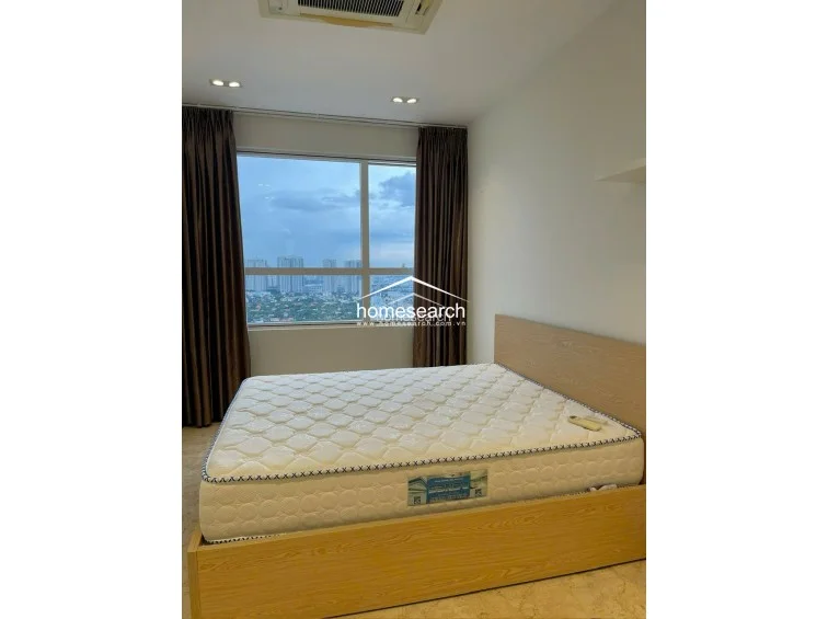 This bedroom is furnished with a light wood-toned bed frame and mattress, complemented by elegant, full-length chocolate-brown curtains. The space maintains a cohesive aesthetic with its light-colored walls, recessed ceiling lighting, and reflective marble flooring.