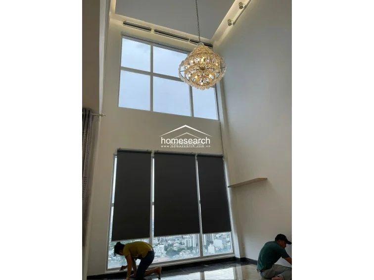 This living space features dramatic double-height ceilings and massive floor-to-ceiling windows that flood the room with natural light. A sophisticated crystal chandelier serves as a focal point, while the neutral walls and dark tile flooring provide a contemporary, high-end feel.
