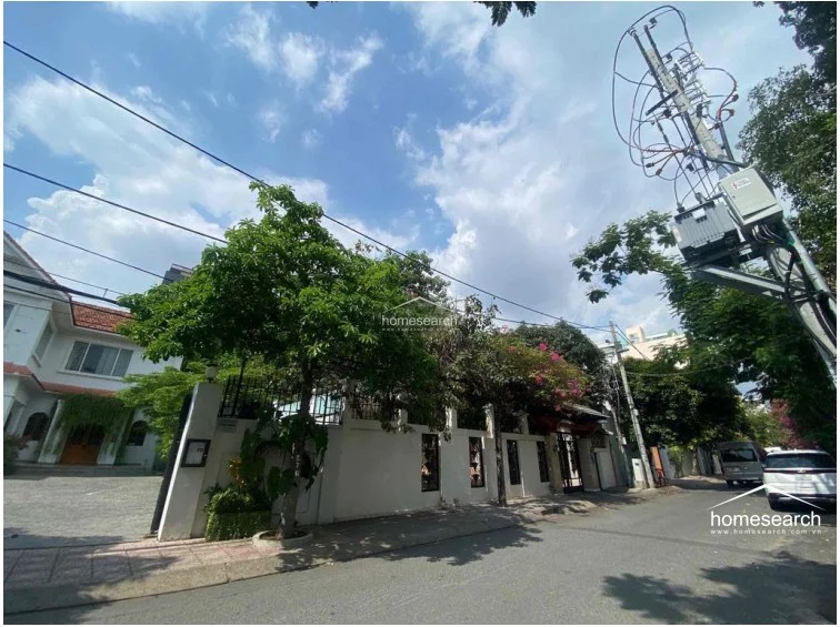 This image shows the exterior street view of a white, multi-story villa featuring a gated entrance and a paved driveway. The property is surrounded by mature trees and lush greenery, providing a sense of privacy and established landscaping.