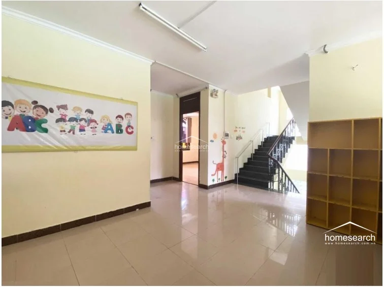 This image shows a spacious landing or common area featuring glossy tile flooring and light yellow walls decorated with children's educational murals. A prominent staircase with a metal railing connects the levels, while an open wooden shelving unit provides functional storage for books or toys.